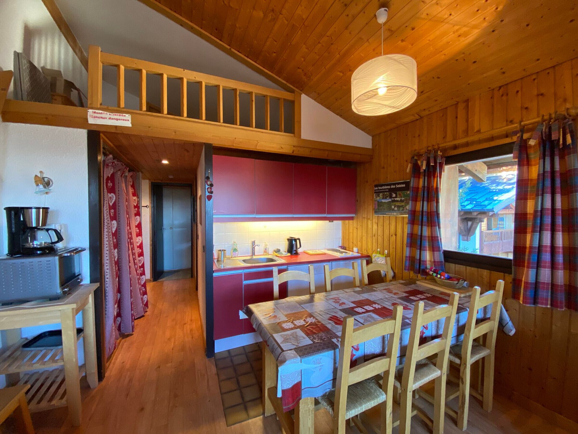Een gezellig cabin interieur met een houten plafond, een eettafel met stoelen, een keuken met rode kastjes en een raam met ruitersgordijnen.