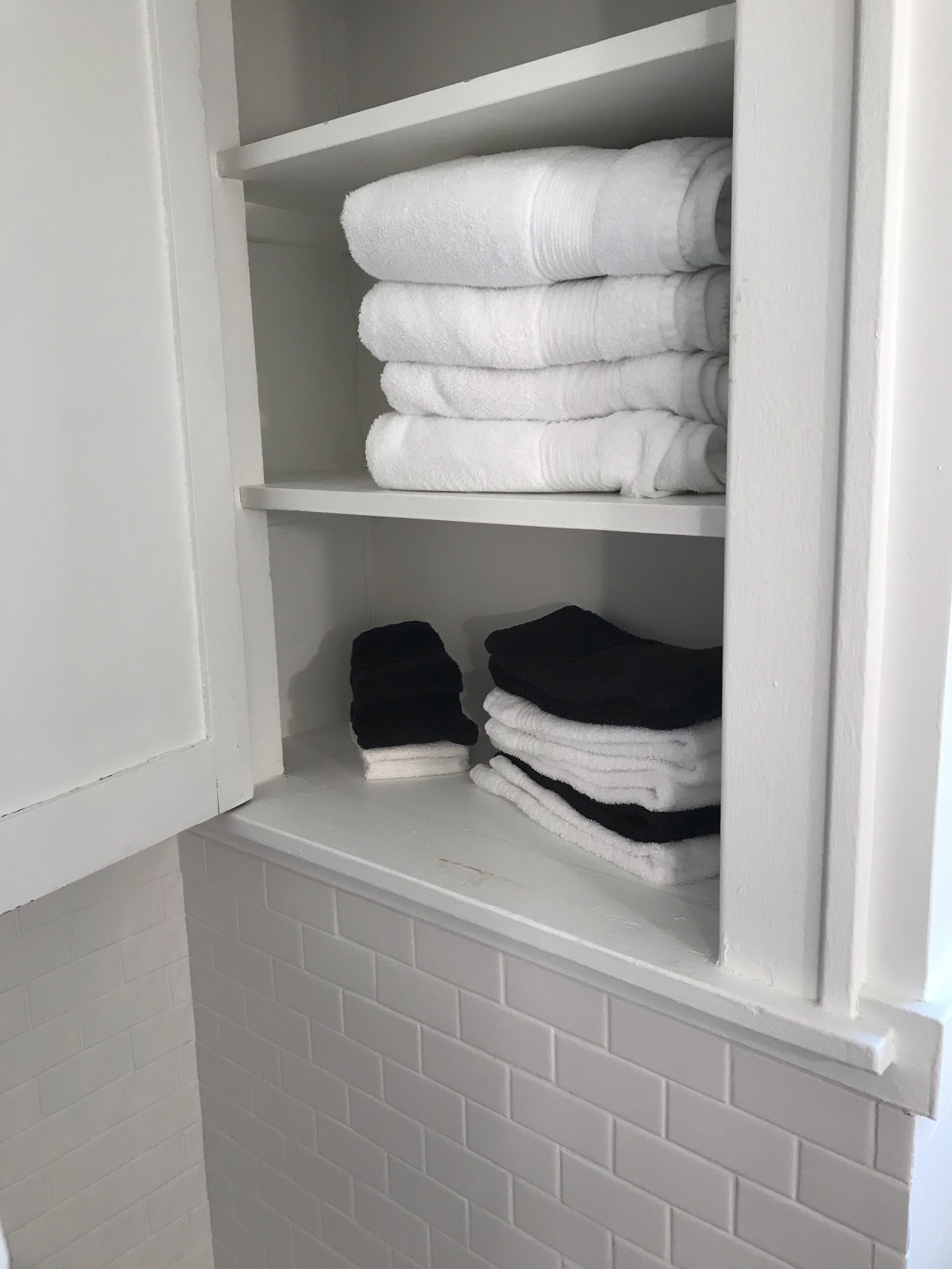 A white tiled bathroom with a white towel shelf containing neatly folded white and black towels.