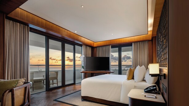 A modern hotel room with a large bed, a flat-screen TV, a bedside table, and a view of the sunset through floor-to-ceiling windows. - Inna Bali Beach Garden (Denpasar)