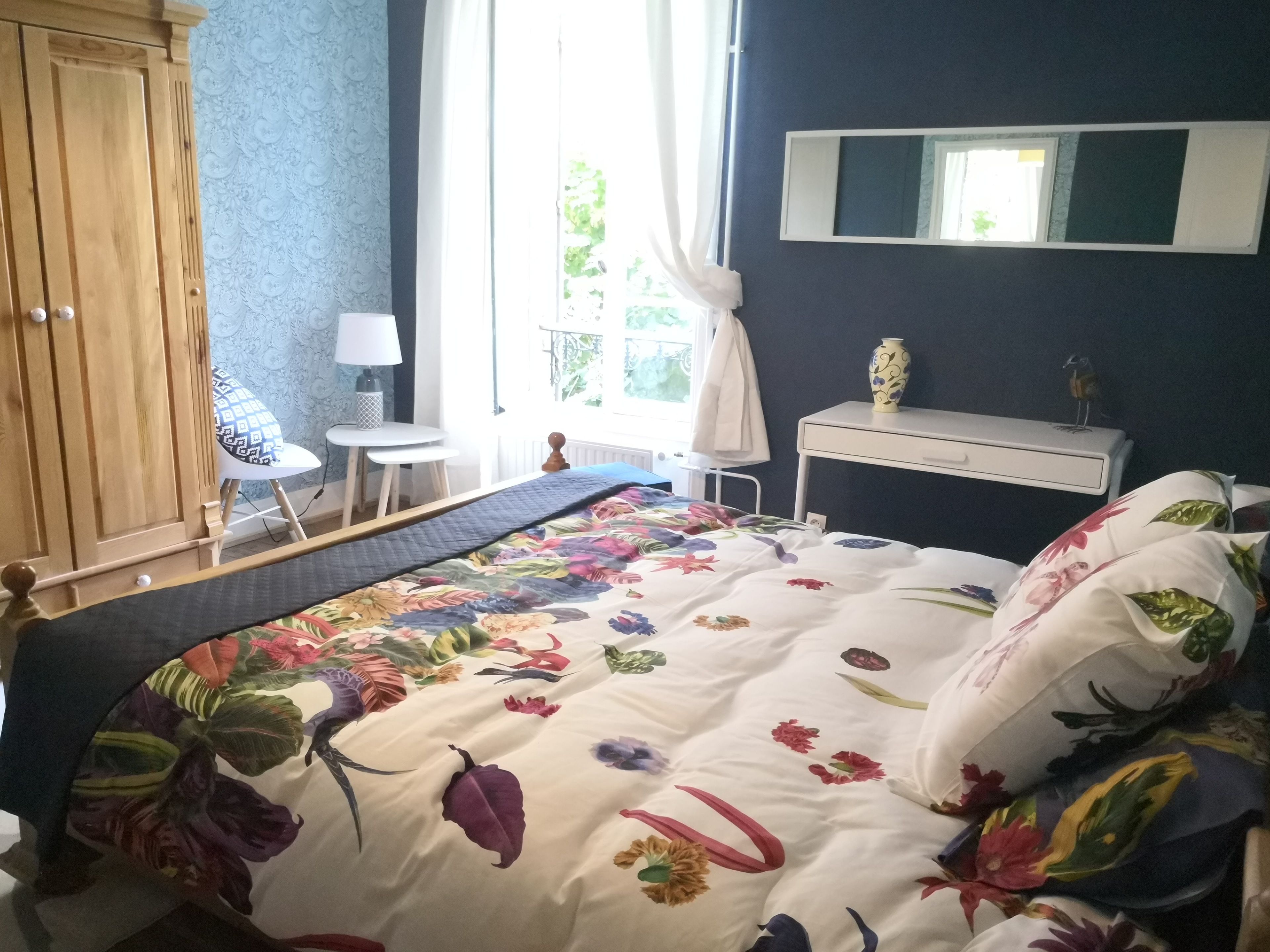 A bedroom with a floral bedspread, a wooden wardrobe, a small white desk, a chair, a bedside table with a vase, and a window with curtains.