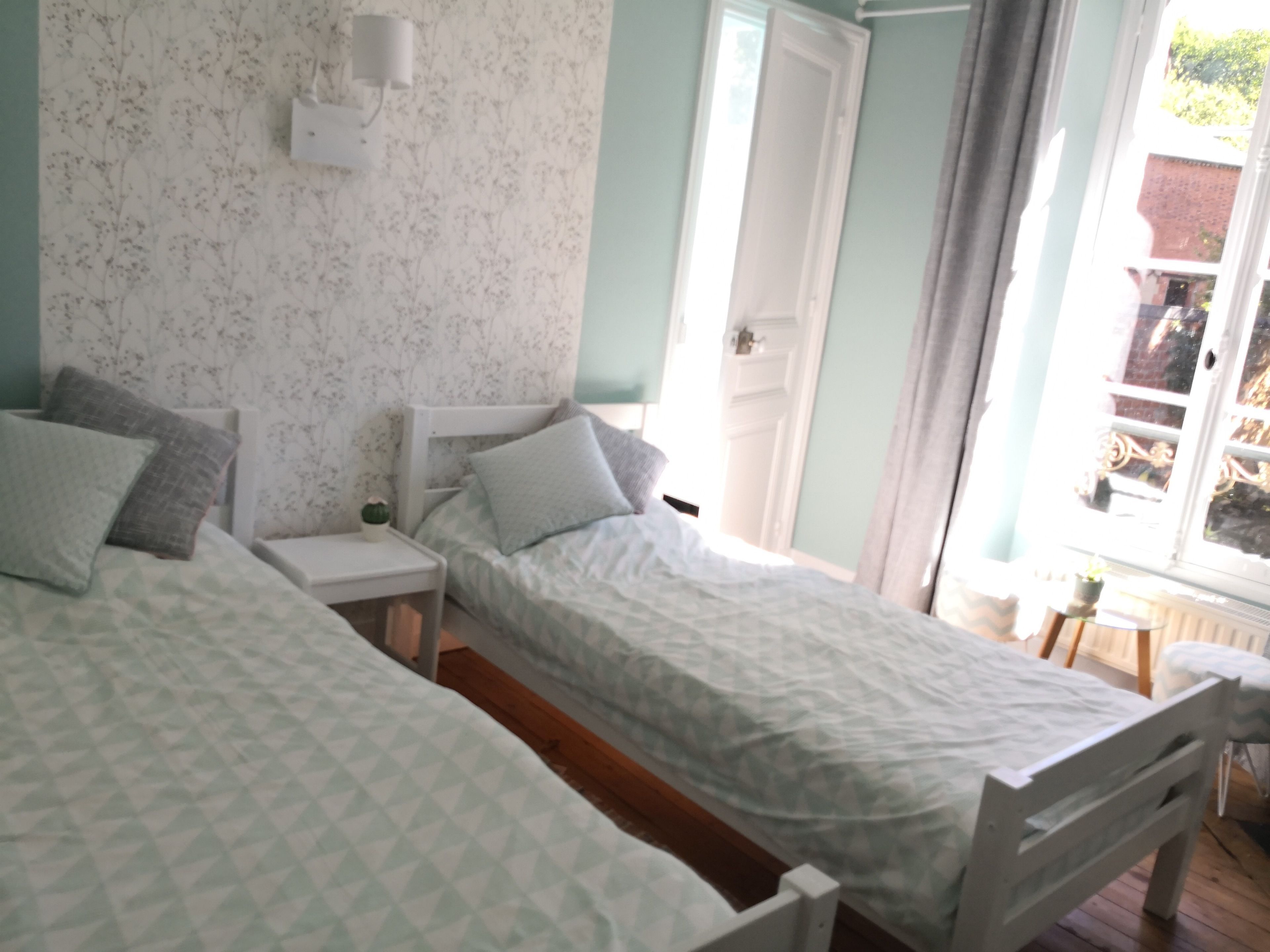 Une chambre avec deux lits simples, une table de chevet, une lampe fixée au mur et une fenêtre avec des rideaux.