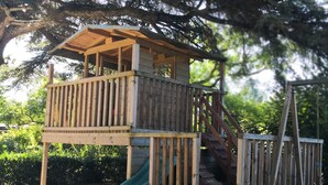 Una casita de juegos de madera con un tobogán, rodeada de árboles y un suelo arenoso.