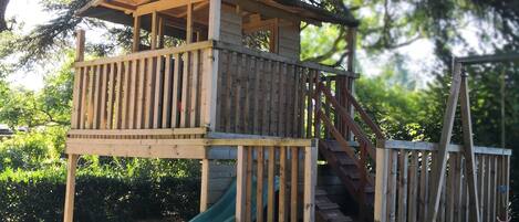 A wooden playhouse with a slide, surrounded by trees and a sandy ground.