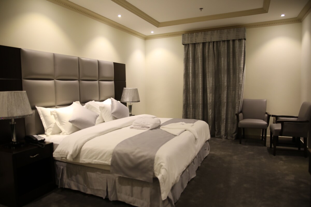 Quarto de hotel com uma cama grande, mesa de cabeceira com luminária, uma cadeira e janela com cortinas.