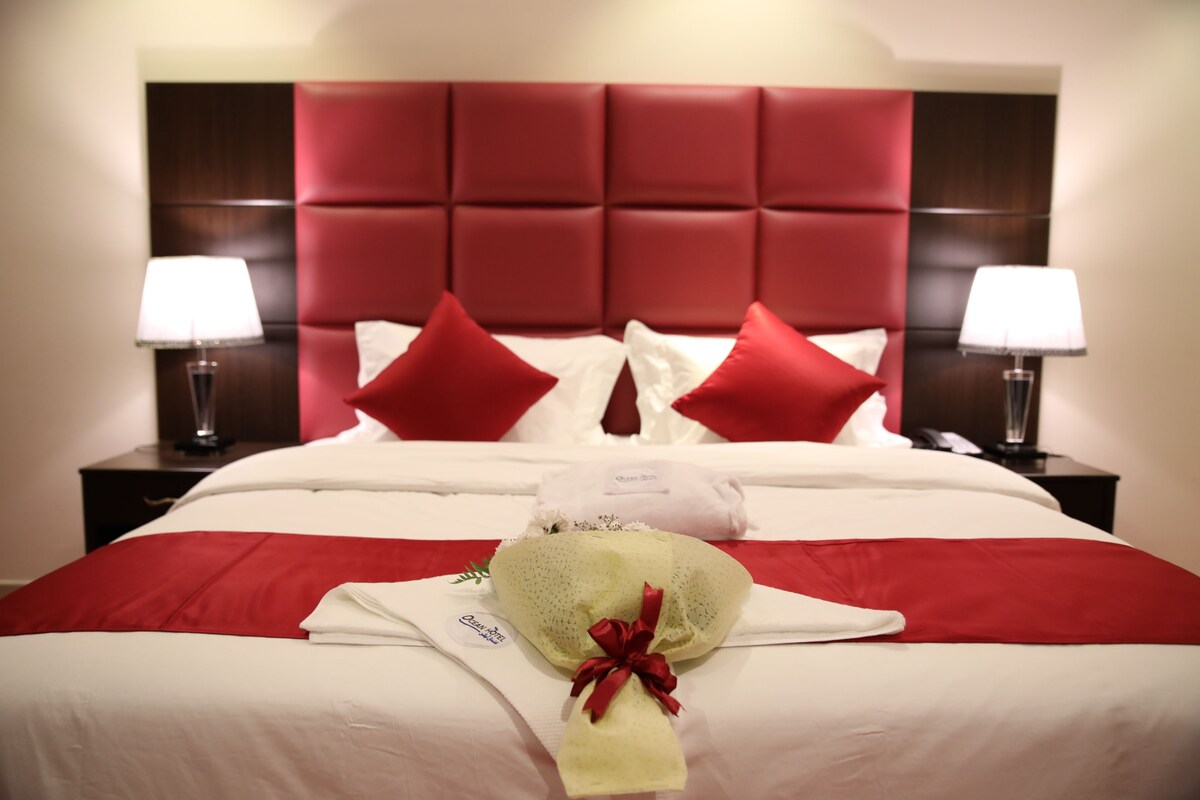 Quarto de hotel com cabeceira estofada em vermelho, roupa de cama branca e um arranjo de flores sobre a cama.