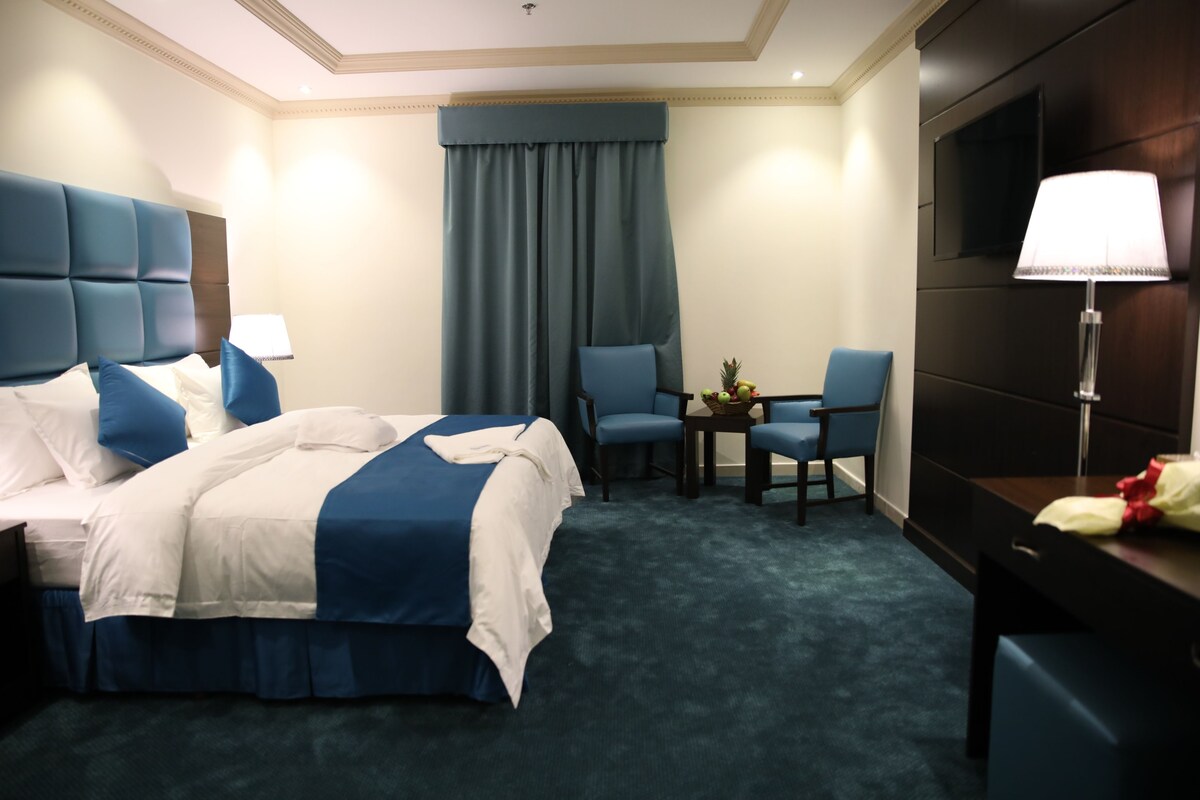Quarto de hotel com uma cama grande, uma escrivaninha, duas cadeiras, uma televisão e um abajur.