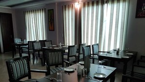 A dining area with multiple tables and chairs, arranged in a spacious room with large windows and vertical blinds. - Mira International (New Delhi)