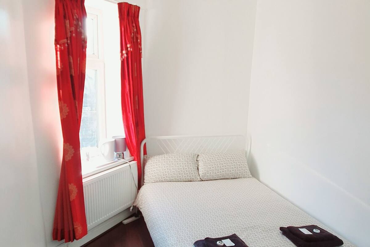 Une petite chambre bien éclairée, avec un lit, un radiateur et un téléviseur.