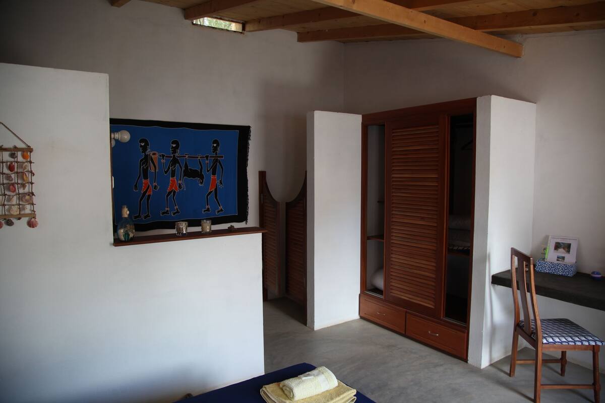 Een kamer met een houten plafond, een kunstwerk in een lijst aan de muur, een houten kledingkast, een stoel en een klein tafeltje met een handdoek erop.
