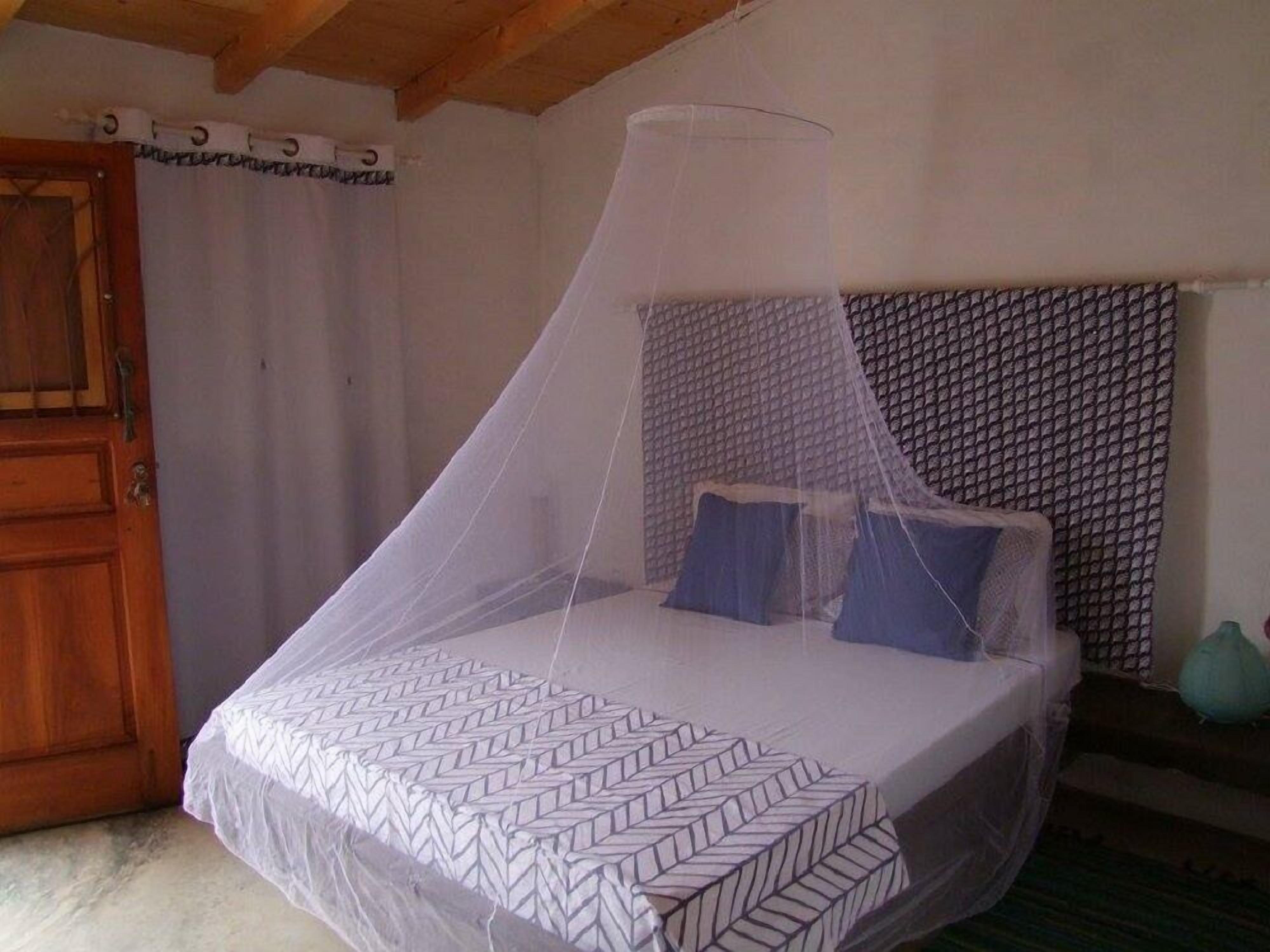 Un lit simple avec un couvre-lit blanc et une moustiquaire, une porte en bois et un mur orné de motifs.