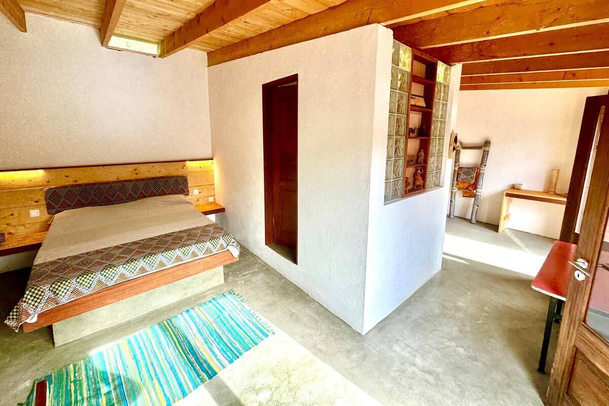 Een slaapkamer met een houten plafond, een bed met beddengoed met een patroon, een klein raam en een houten kast.