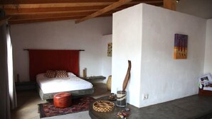 Une chambre à coucher avec un lit, un rideau rouge, un plafond avec une poutre en bois, une petite table avec un verre et un objet en bois, et un mur orné d’un tableau.
