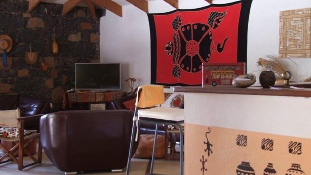 Un salon chaleureux avec un mur en pierre, des poutres apparentes, un téléviseur à écran plat, un canapé en cuir marron, une table basse en bois et un objet décoratif rouge.