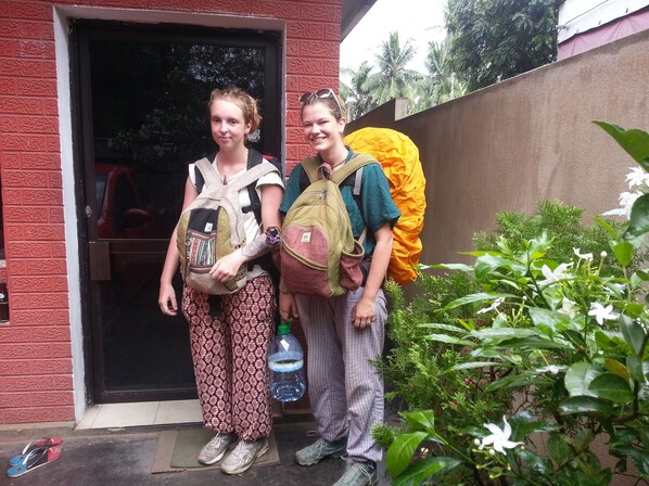 Two individuals standing at a doorway with backpacks, a water bottle, and flip-flops on the ground.