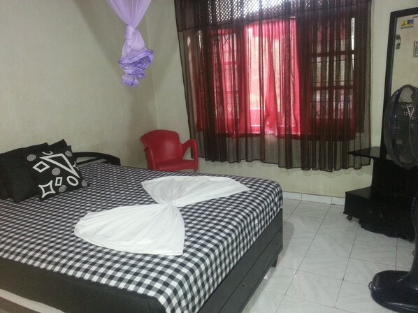 A hotel room with a checkered bedspread, a red chair, a black fan, and a window with red curtains.