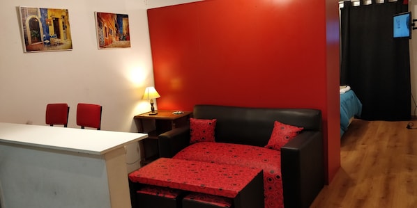 A room with a red wall, a bed with a red patterned bedspread, a desk with two red chairs, and a small table with a lamp.