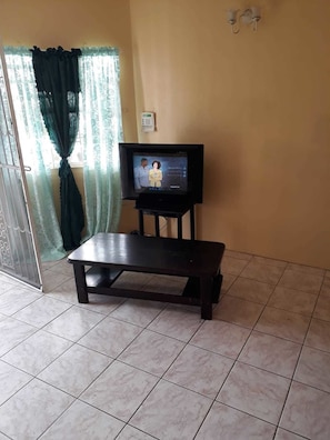 Ein Zimmer mit Fernseher auf einem Schrank, einem kleinen Tisch, einem Vorhang und einem gefliesten Boden.