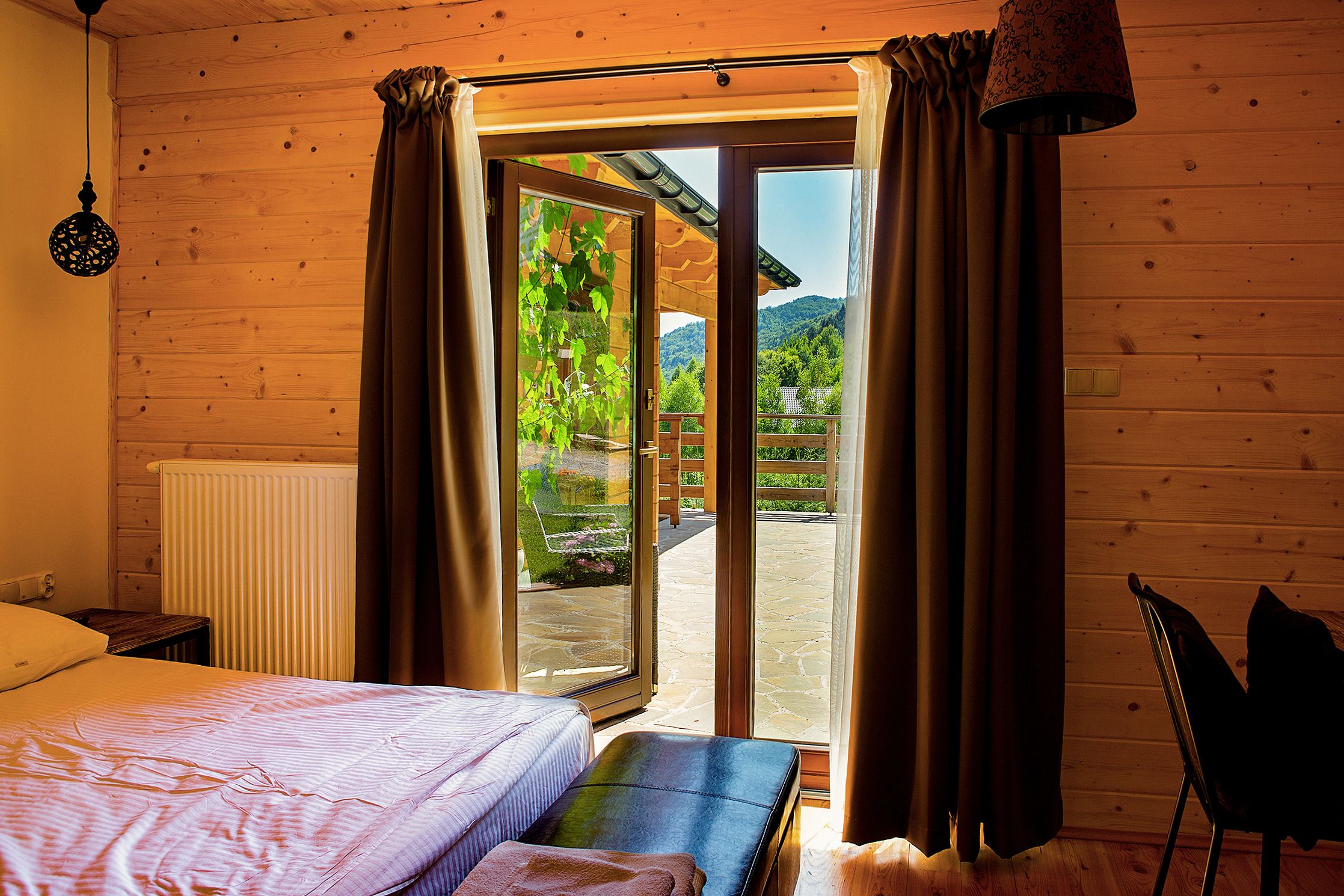 Quarto de cabana de madeira com uma cama, um banco e vista para um jardim e montanhas por uma porta de vidro.