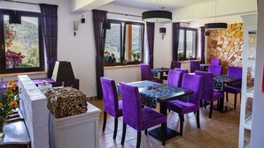 A dining area with purple chairs and tables, large windows with curtains, and a stone wall.