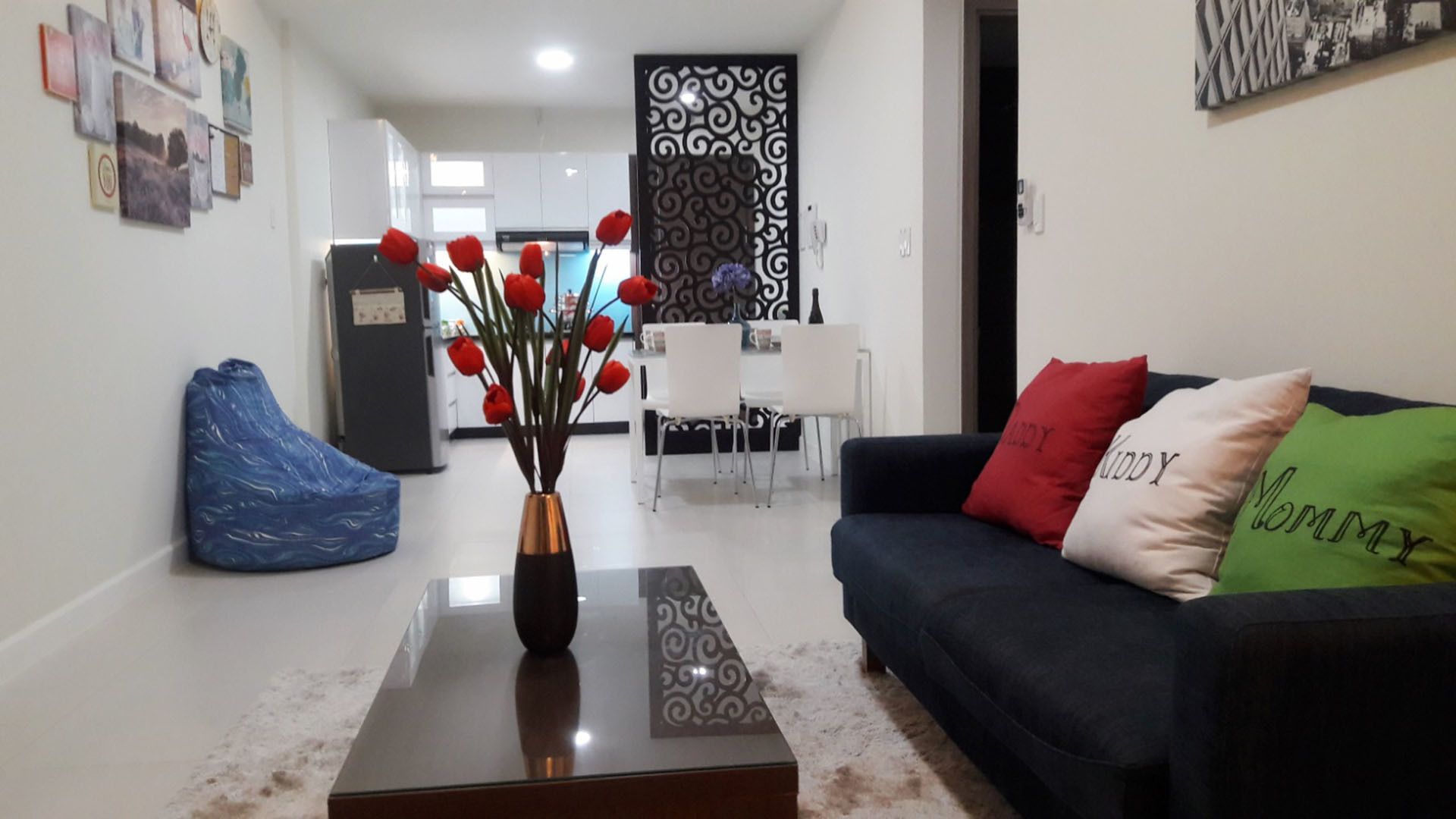 A living room with a coffee table, a sofa with decorative pillows, a blue bean bag chair, and a dining area in the background.
