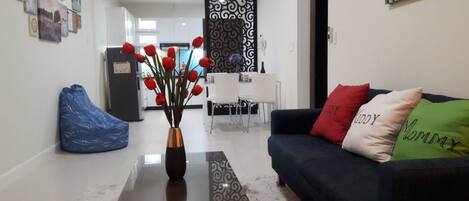 A living room with a coffee table, a sofa with decorative pillows, a blue bean bag chair, and a dining area in the background.