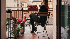 A person sitting on a wooden chair on a balcony with a table and potted plants.