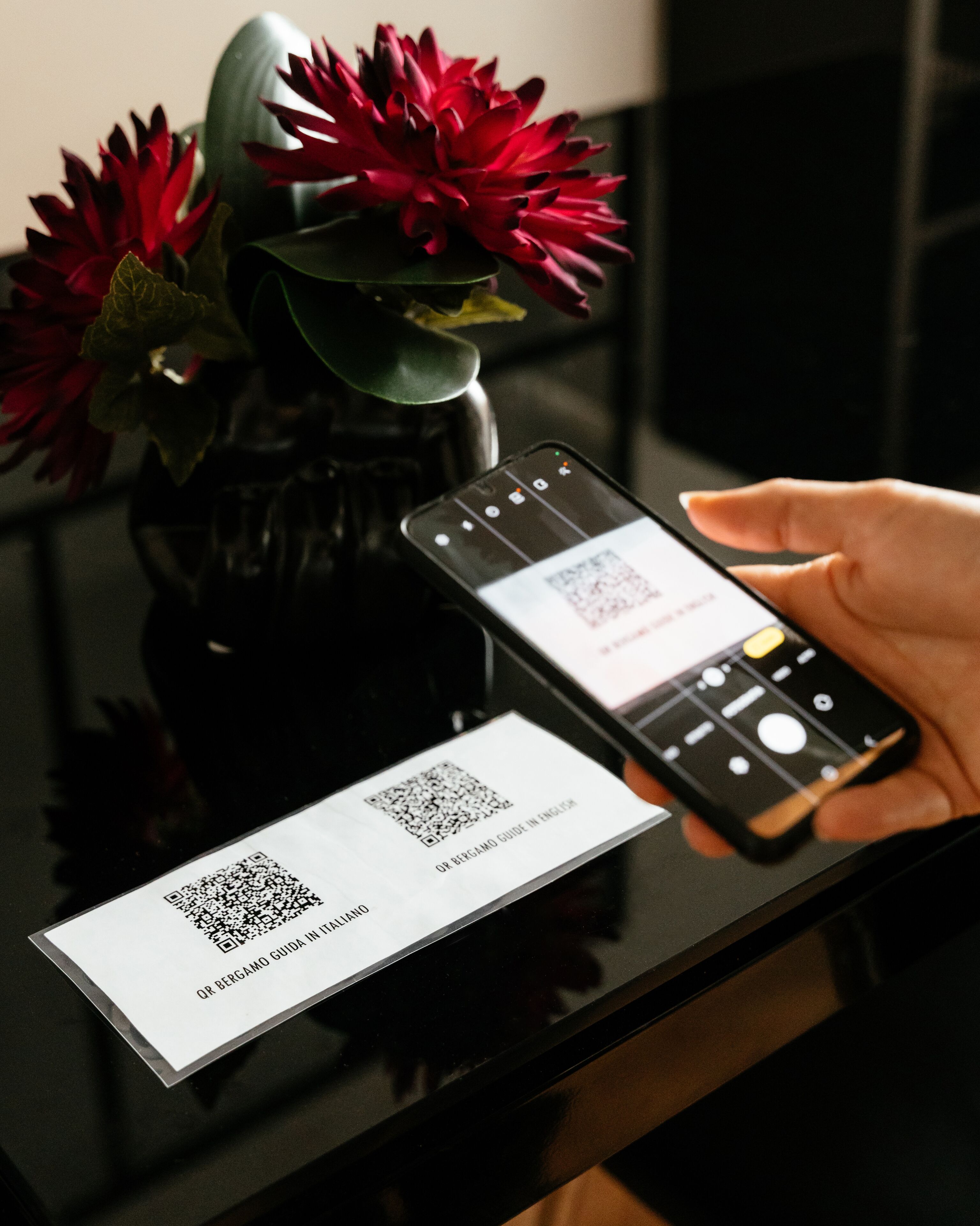 A hand holding a smartphone with a QR code and a business card on a reflective surface.