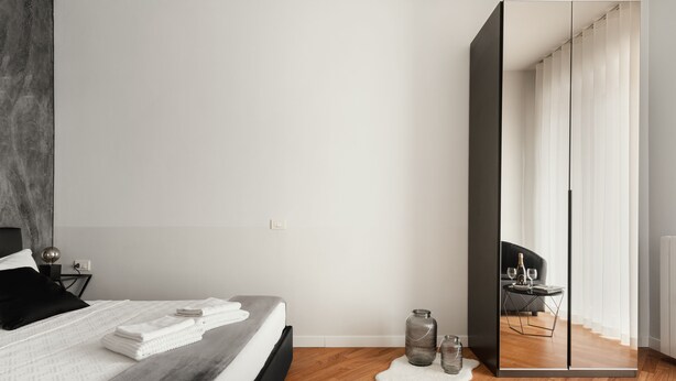 A modern bedroom with a large bed, a bedside table with a lamp, and a mirror reflecting a living area.