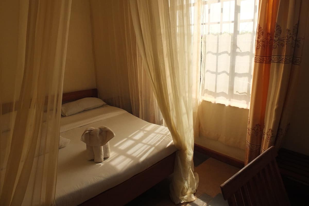 Una cama individual con dosel, un peluche pequeño y una ventana con cortinas traslúcidas.