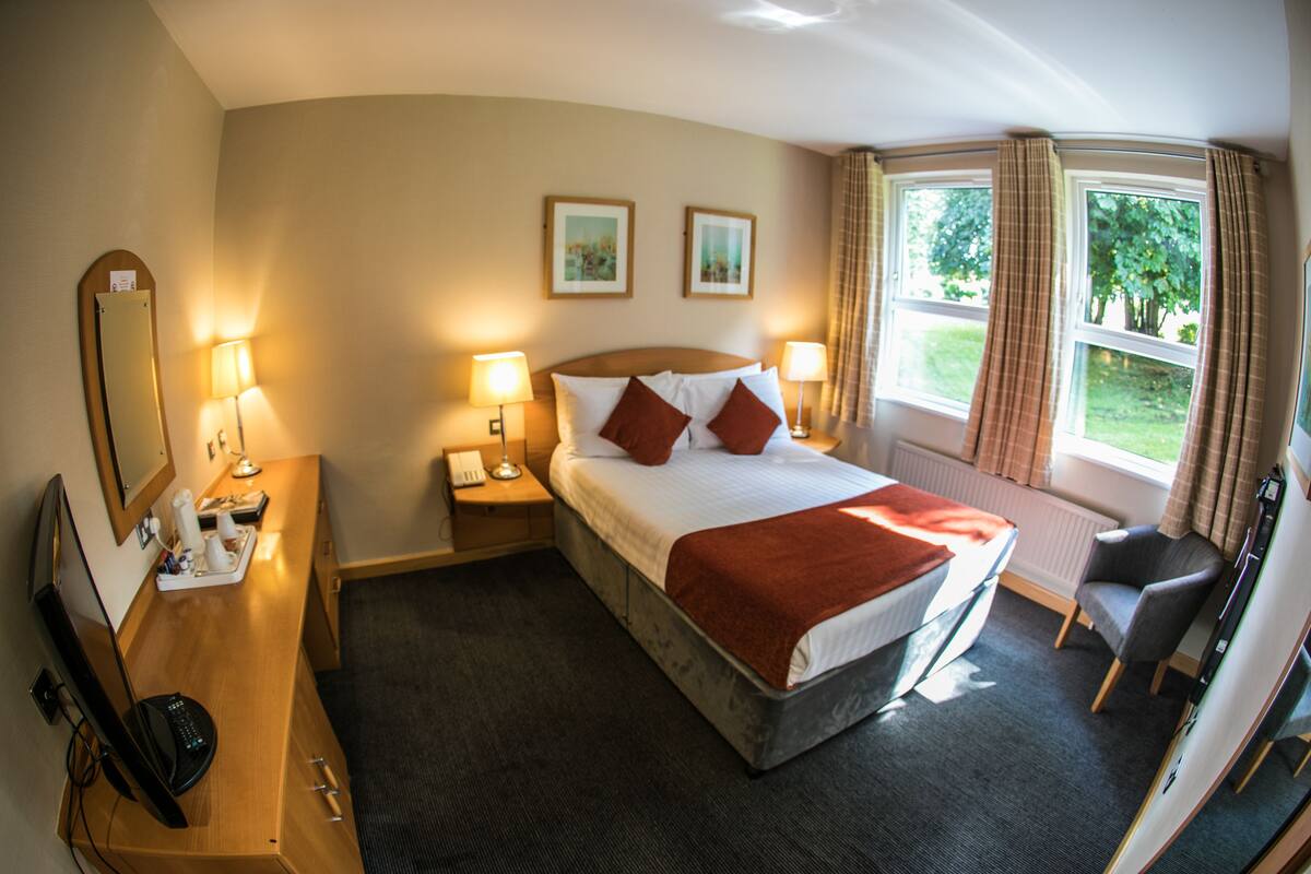 A hotel room with a bed, two bedside lamps, a desk with a computer, a chair, and a window with curtains.