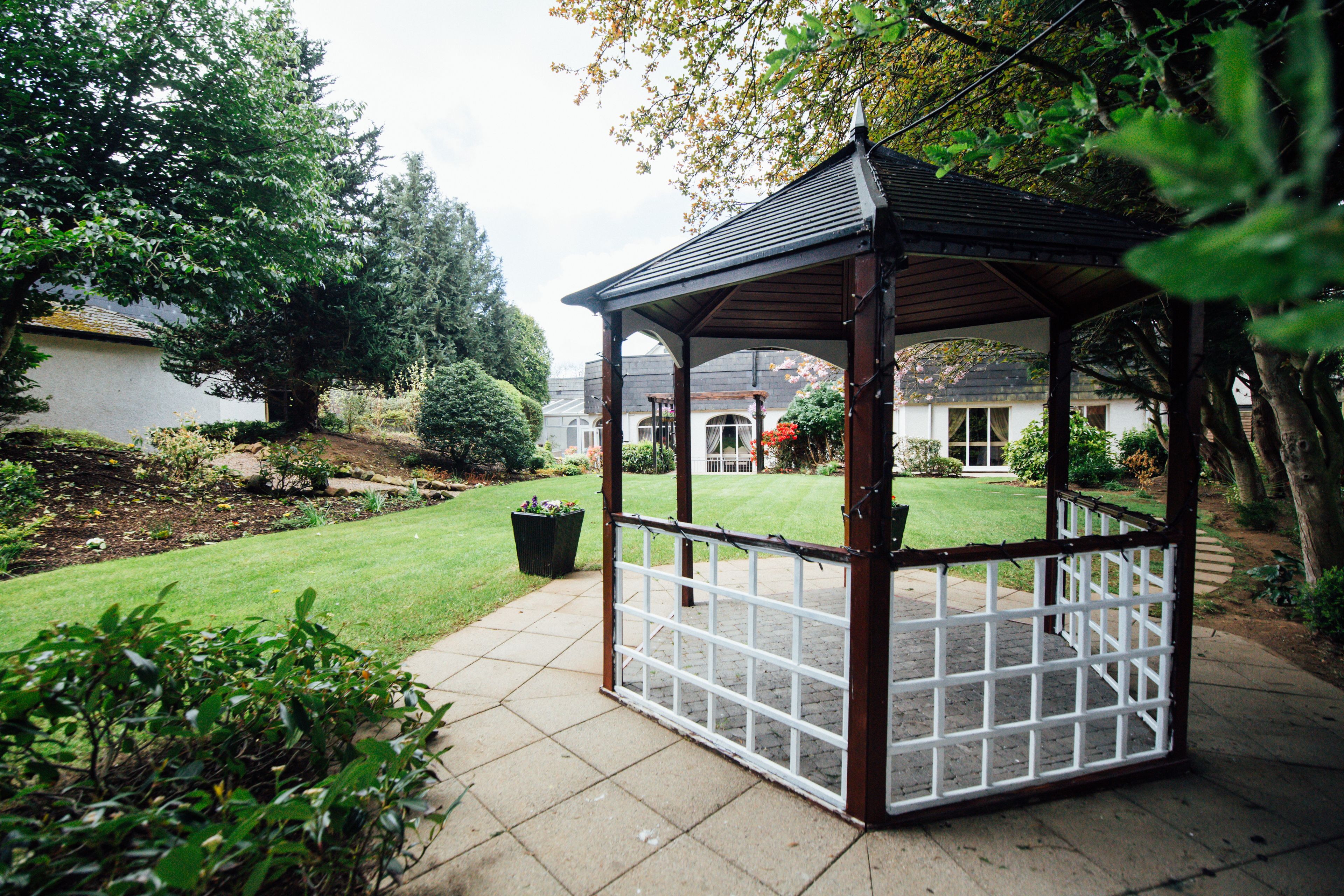 A gazebo with a tiled floor and a wooden frame, surrounded by a well-maintained garden and a lawn.