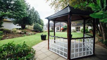 Un gazebo con pavimento in piastrelle e struttura in legno, immerso in un giardino curato e un prato.