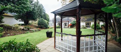 A gazebo with a tiled floor and a wooden frame, surrounded by a well-maintained garden and a lawn.