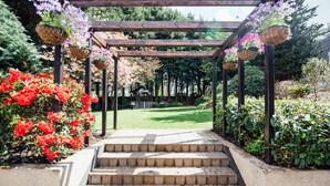 A wooden pergola with hanging baskets of flowers, a paved pathway, and a garden with various plants and a gazebo in the background. - Rosspark Hotel (Ballymena)