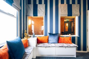 Una habitación con pared azul y blanca a rayas, una ventana con cortinas, un banco con cojines y una estatua en el alféizar.