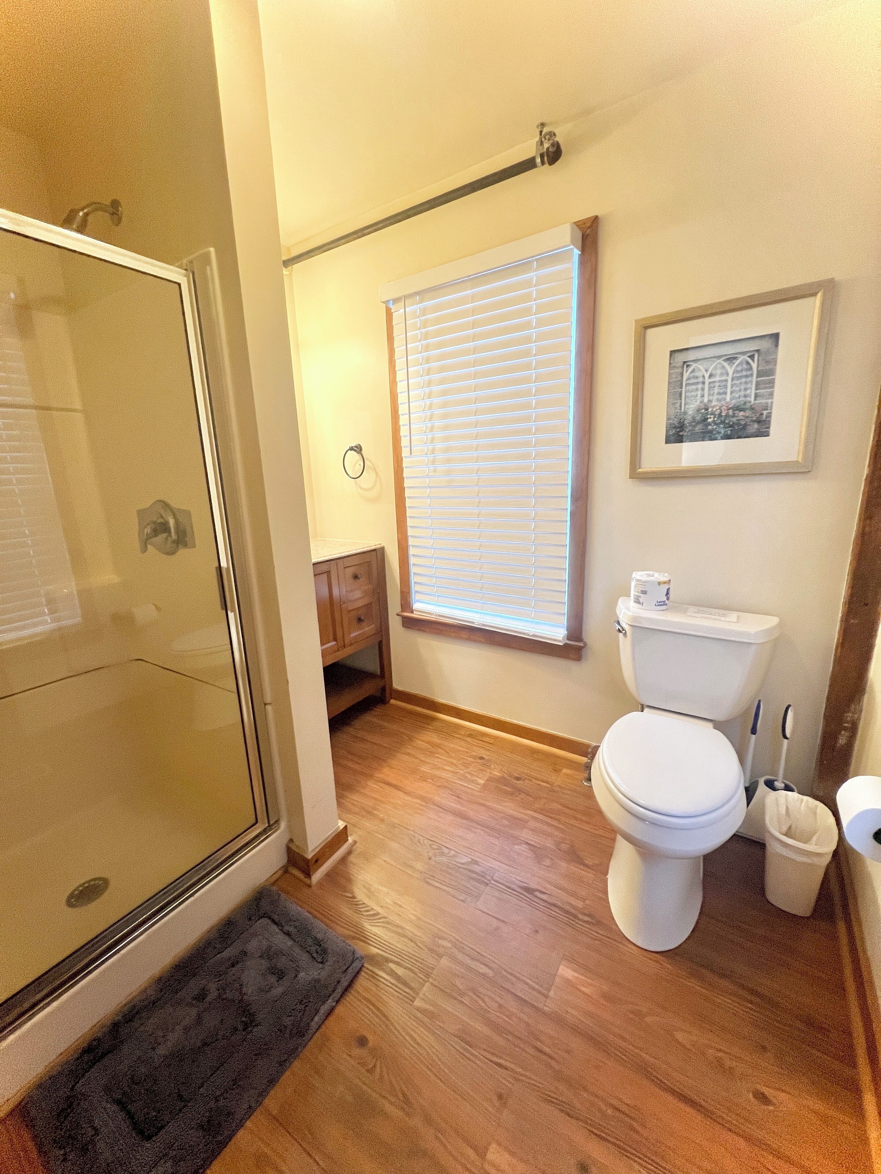 Une salle de bain avec une douche à parois de verre, des toilettes, une fenêtre avec des stores, un tableau encadré au mur et un plancher en bois.