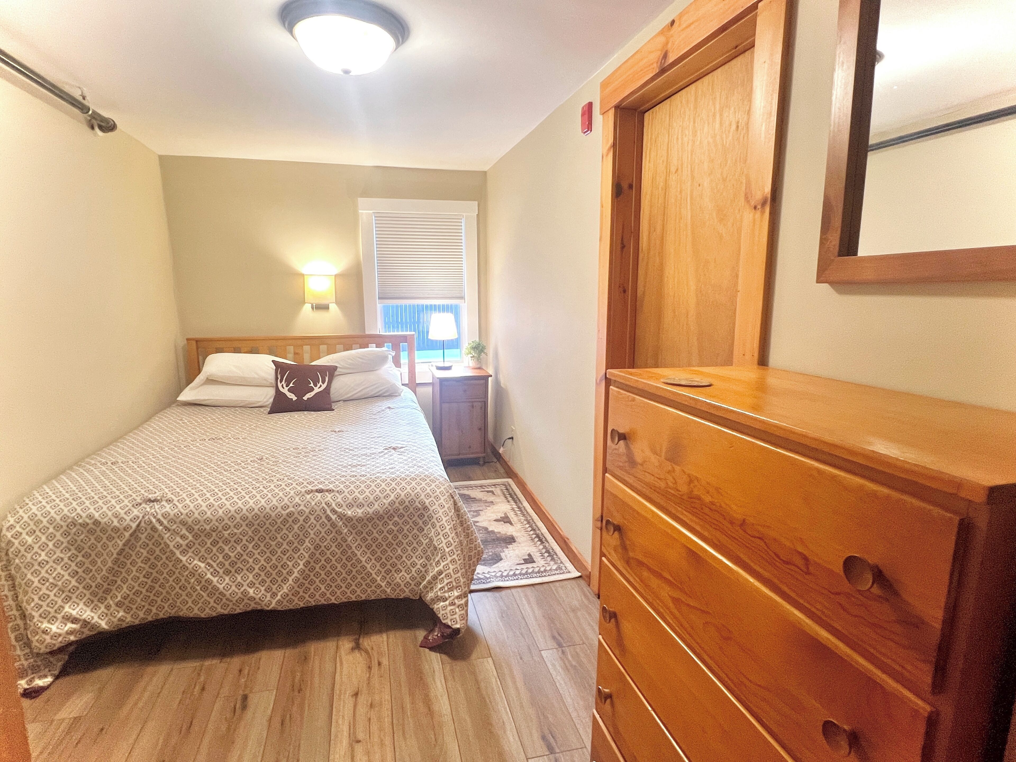 A bedroom with a bed, a dresser, a nightstand, a lamp, and a window with blinds.