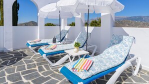 A sunlit outdoor lounge area with white and blue lounge chairs, white umbrellas, and a stone floor. - Moorish-Style Mountain Villa: Stunning 360° Views, Pool & Hot Tub (Torrox)