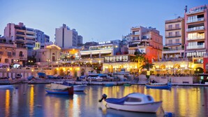 Zona costera con barcos amarrados, un edificio con el letrero 'DOLCE VITA' y una hilera de establecimientos iluminados a lo largo del agua.