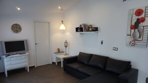 A living room with a black leather sofa, a white TV stand, a small table with a lamp, and a wall-mounted shelf with decorative items. - ATTIC + PRIVATE TERRACE AND FREE WIFI (Sevilla)