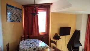 A dining area with a table covered in a patterned tablecloth, wooden chairs, a hanging light fixture, a television mounted on the wall, and a fireplace. - Residential flat (Morzine)