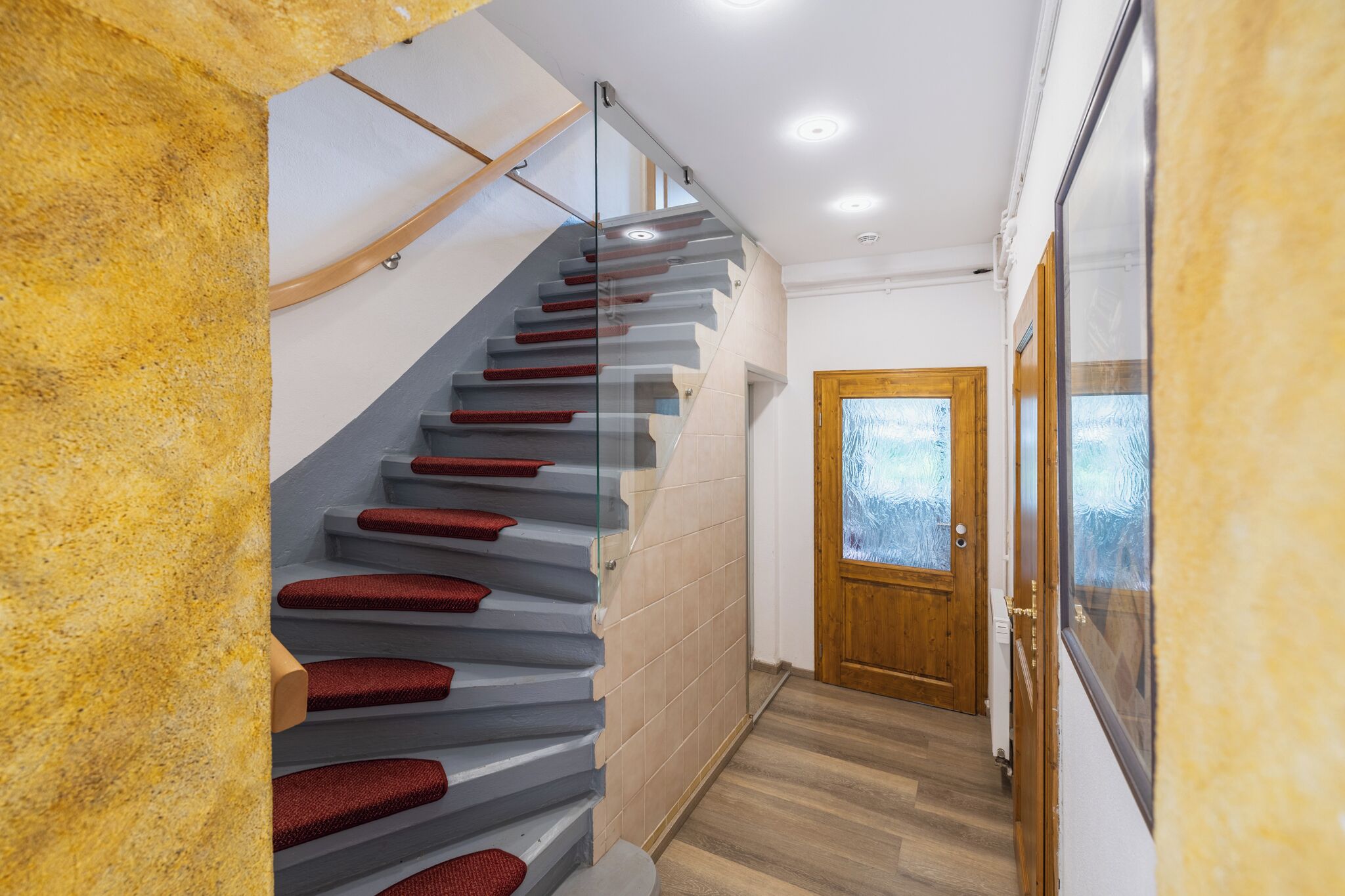 Eine moderne Treppe mit Glastrennfeldern und roten Stufen, die zu einer Holztür mit satinierten Glasscheiben führt.
