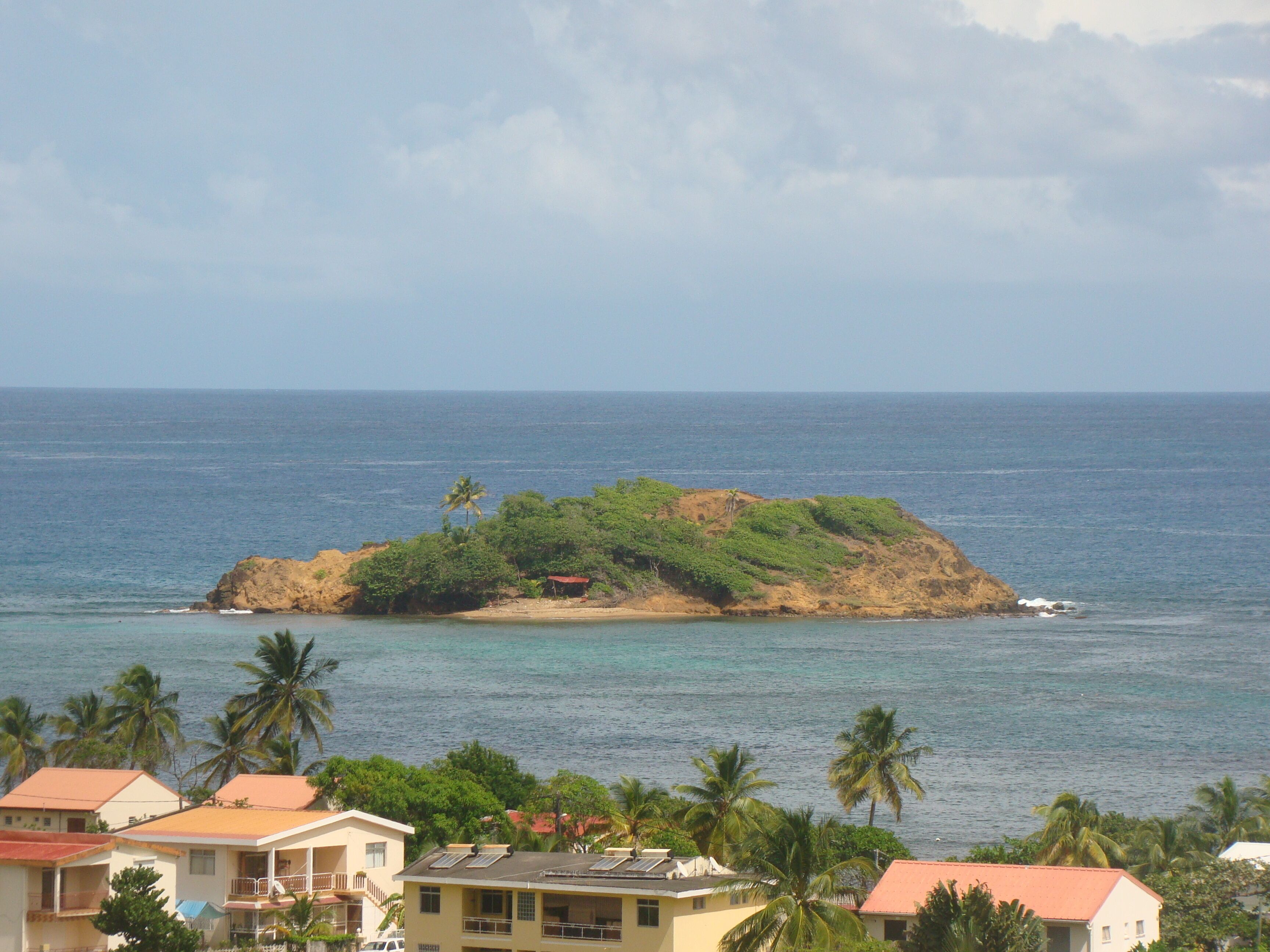 Vista costera con una pequeña isla, casas de techo rojo y palmeras.