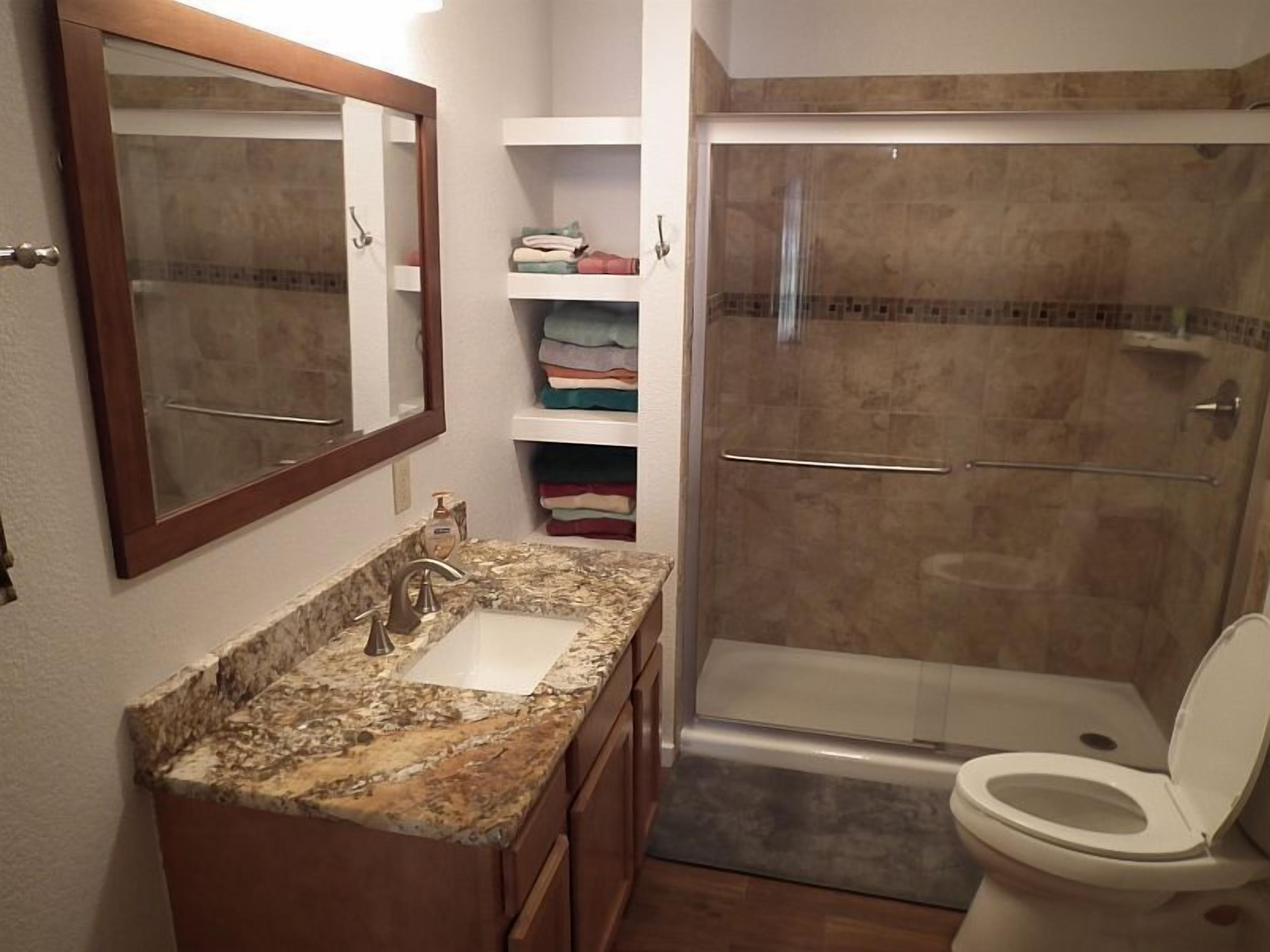 A bathroom with a granite countertop sink, a mirror, a shower enclosure, and a toilet.