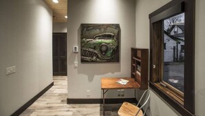 A hallway with a wooden floor, a painting of a vintage car on the wall, a small wooden table with a chair, and a window with a view of a building outside. - Modern Cabin with Walkability in Downtown Driggs (Driggs)