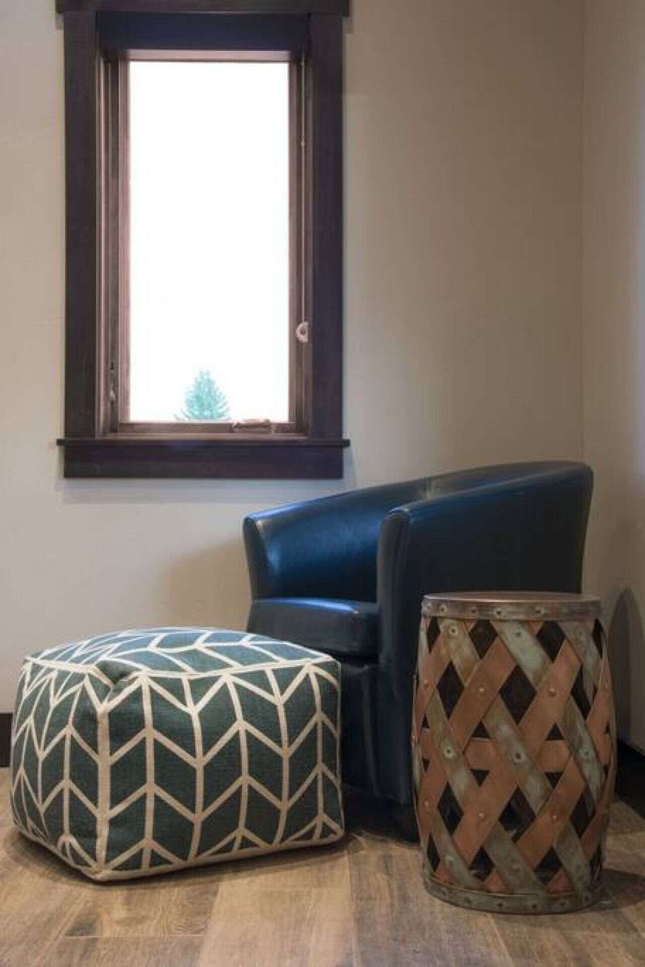Un rincón de una habitación con una ventana, un sillón azul oscuro y dos otomanas con diseños geométricos.