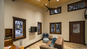 A spacious living room with a wooden ceiling, large windows, a flat-screen TV, a leather chair, and a sofa. - Modern Cabin with Walkability in Downtown Driggs (Driggs)