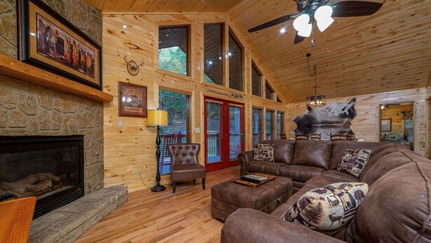 A cozy log cabin interior with a stone fireplace, leather sofas, and a large window overlooking the forest. - Large, Spacious, Relaxing 4 Bedroom 4.5 Bath Pigeon Forge Tennessee Cabin (Sevierville)