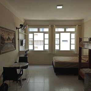 A dormitory room with a bed, a desk, a chair, a fan, and a window with curtains. - Apartamento Estudio na Pitangueiras (Guarujá)