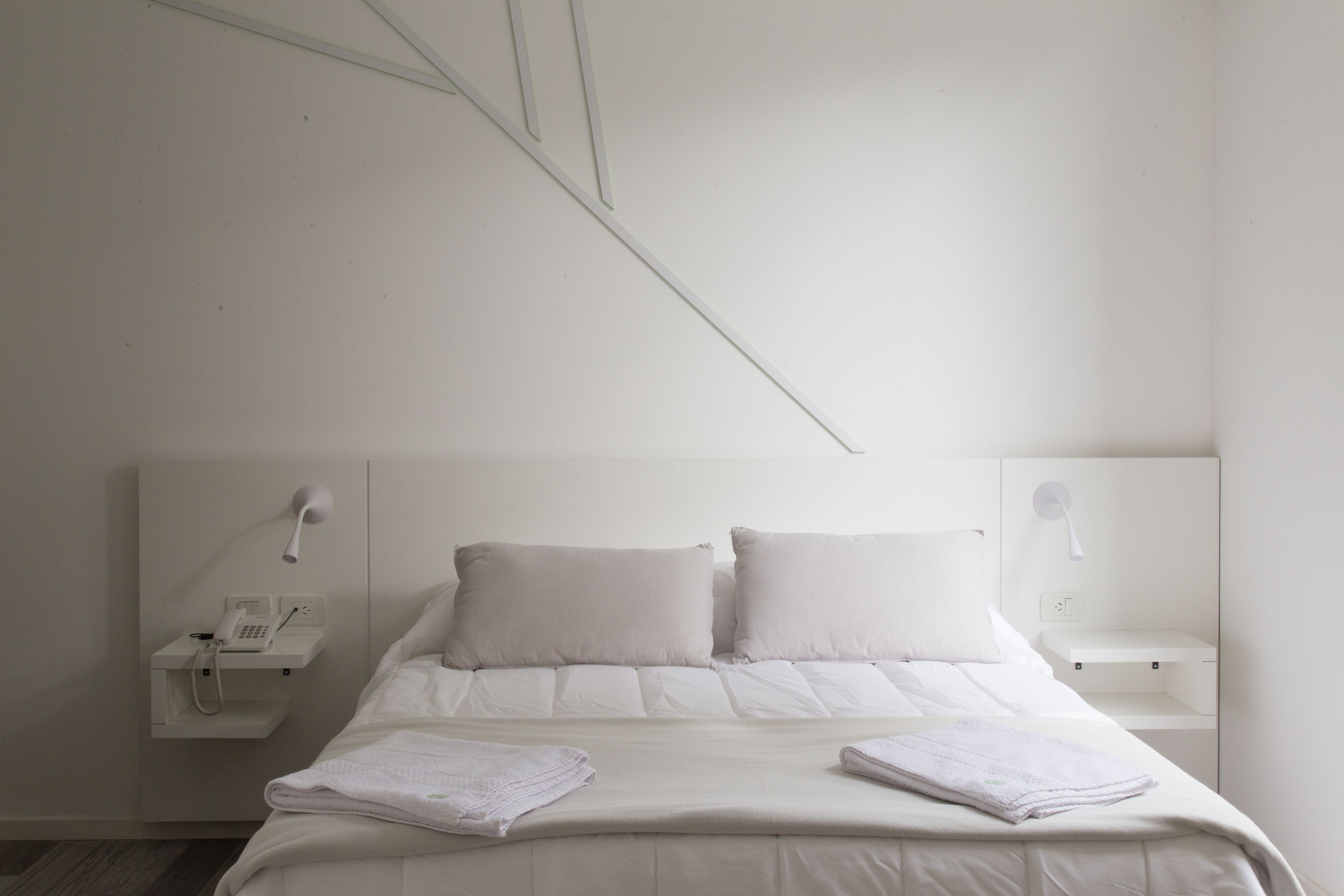 A minimalist hotel room with a bed, two pillows, a bedside table with a lamp, and a phone.